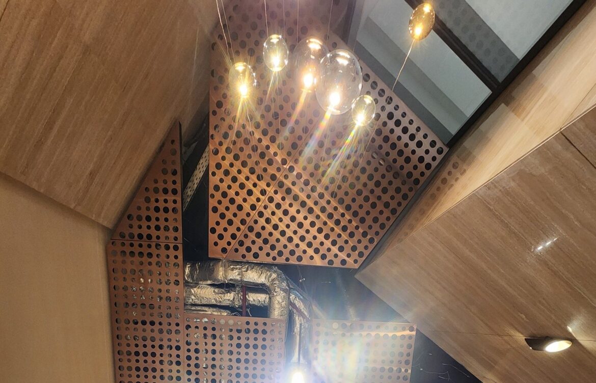 Looking up into a high entrance hall: perforated copper metal ceiling, glass pendants and ducts.