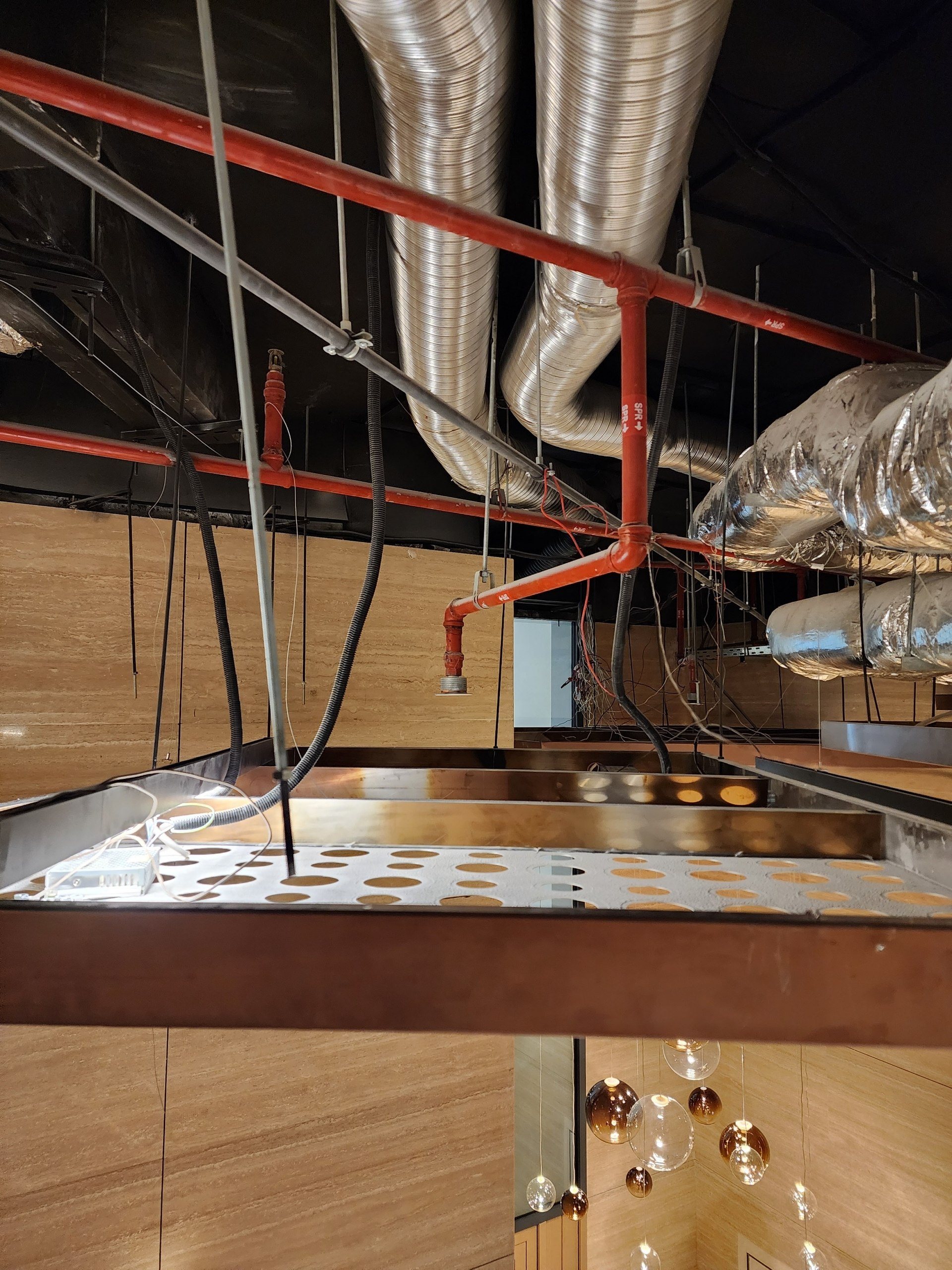 Insulated ducts and sprinkler pipes above an open perforated ceiling panel.