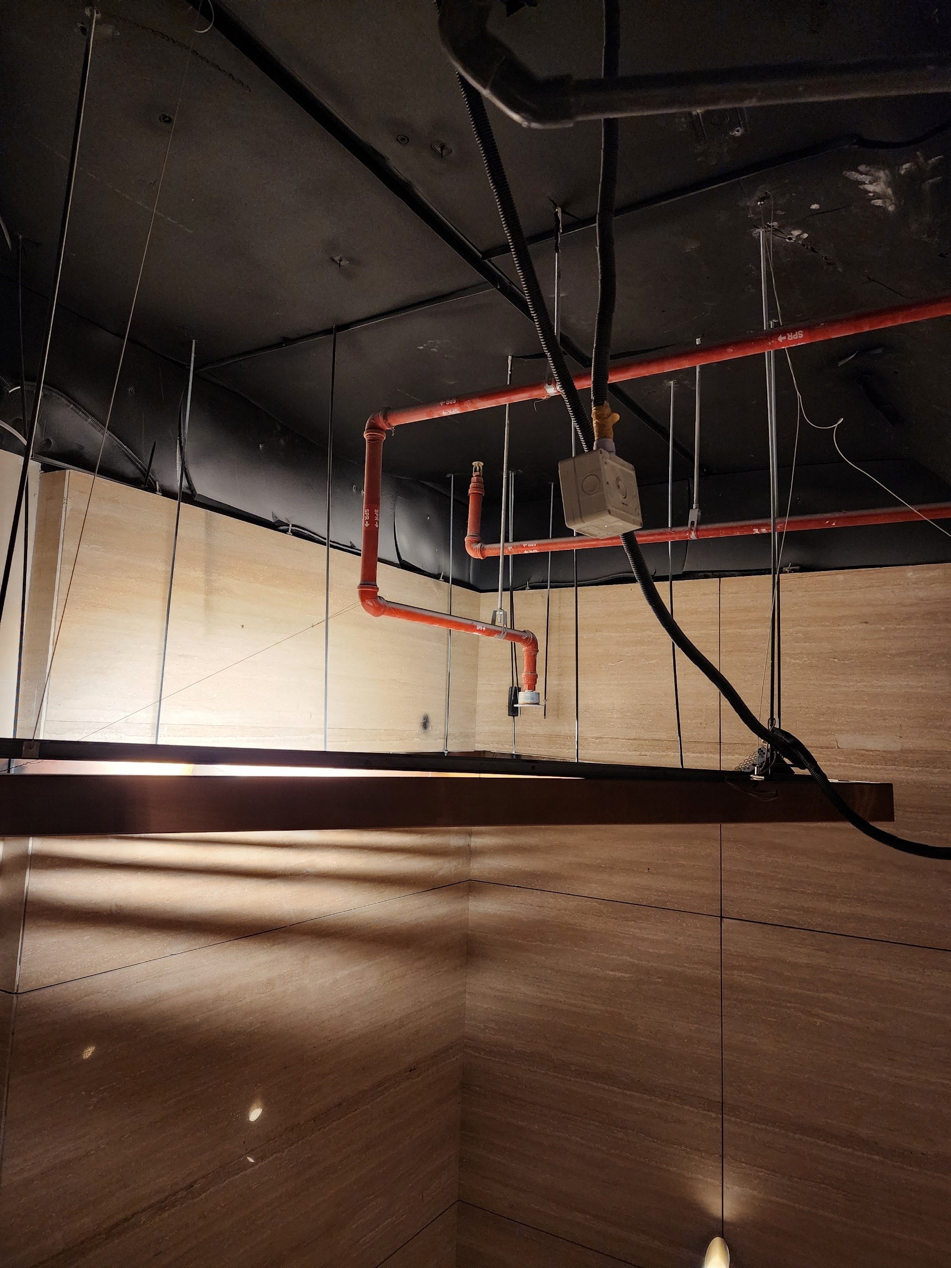 Sprinkler pipes and cables above a hanging metal ceiling panel on travertine walls.