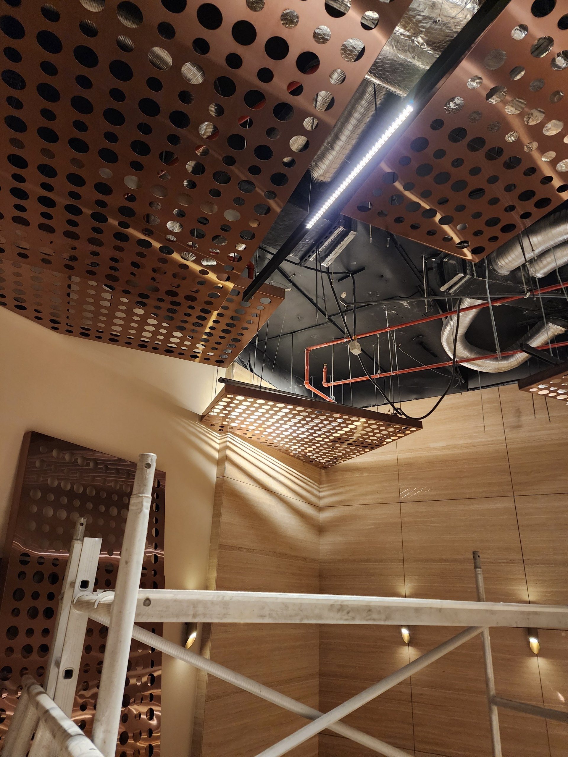 Perforated metal ceiling panels being hung over travertine walls, scaffolding in the hall.
