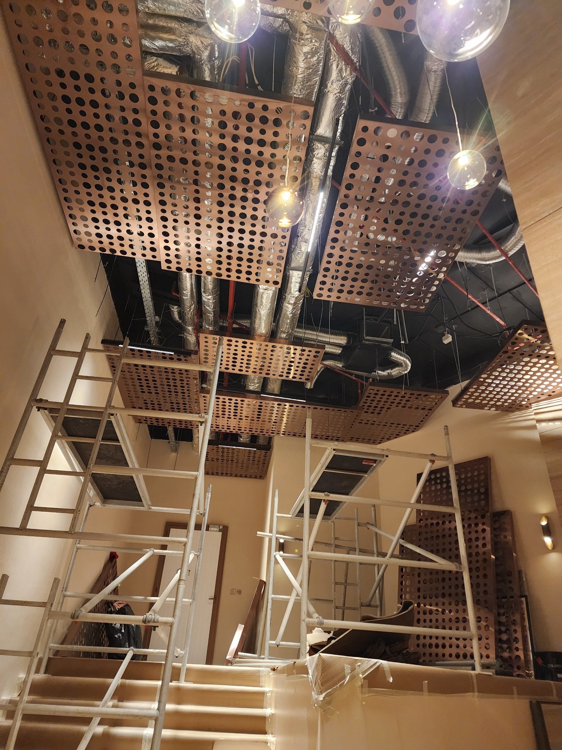The full hall during ceiling install: scaffolding, stepped perforated panels, ducts and glass pendants.